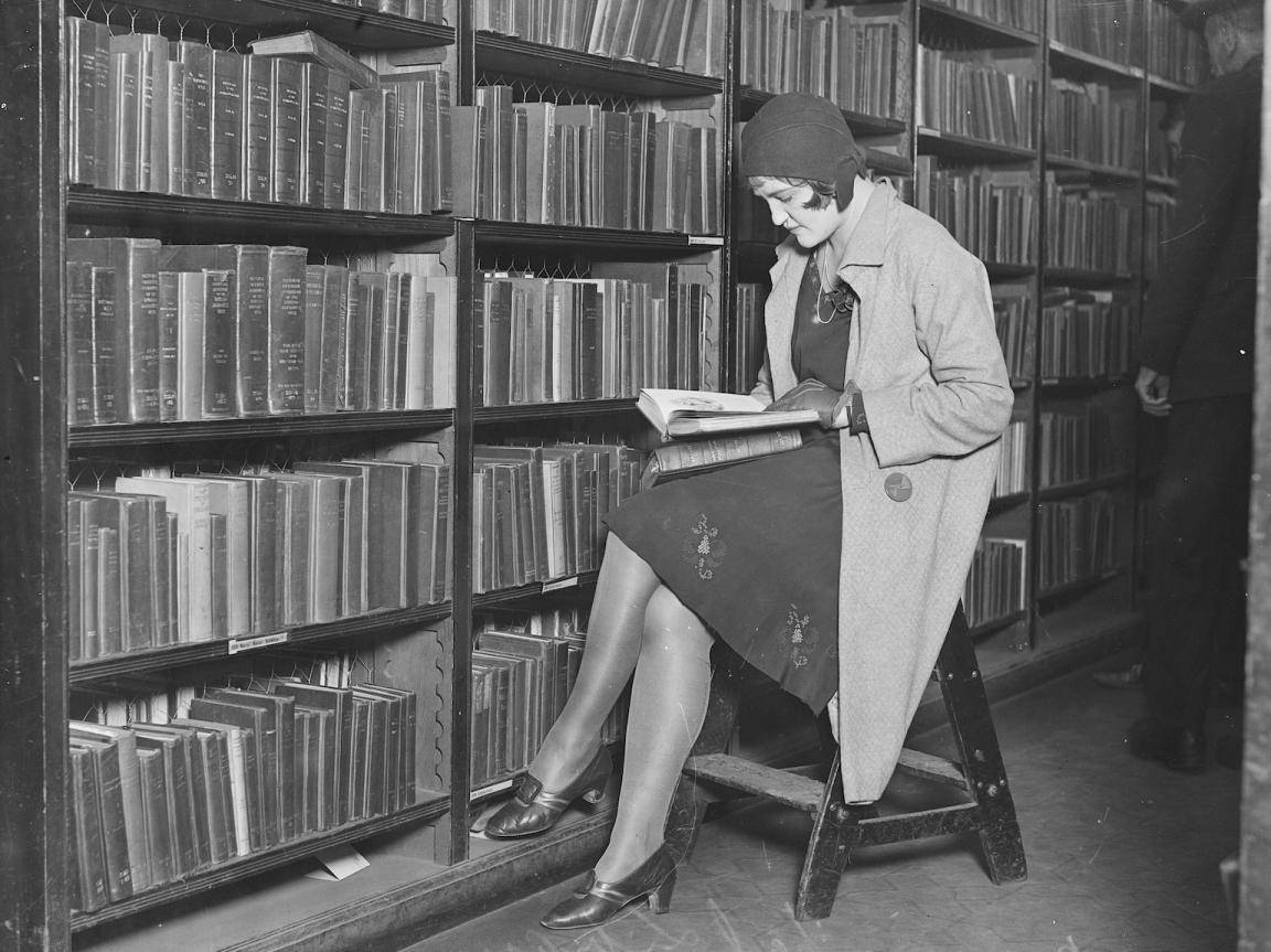 Black and white photo of a woman in winter clothes sitting on a stool by a bookshelf in the library reading a book