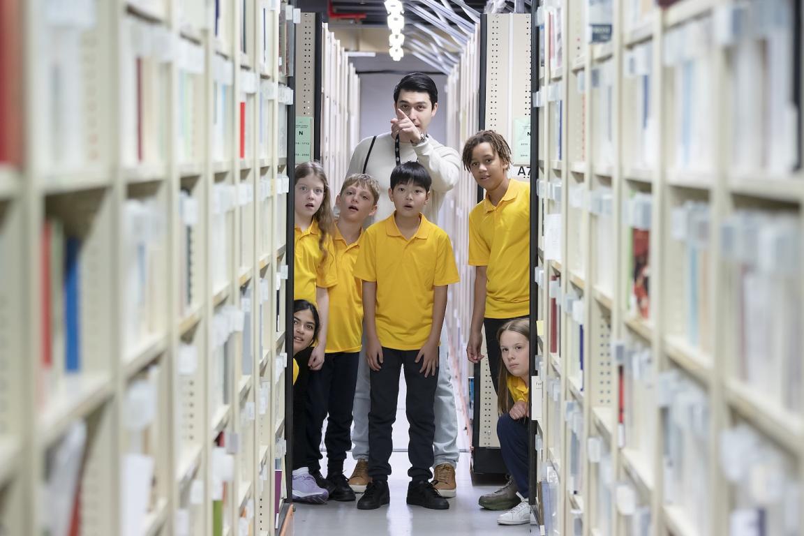 Six young school children standing in the stacks with a Library staff member look at material on shelves