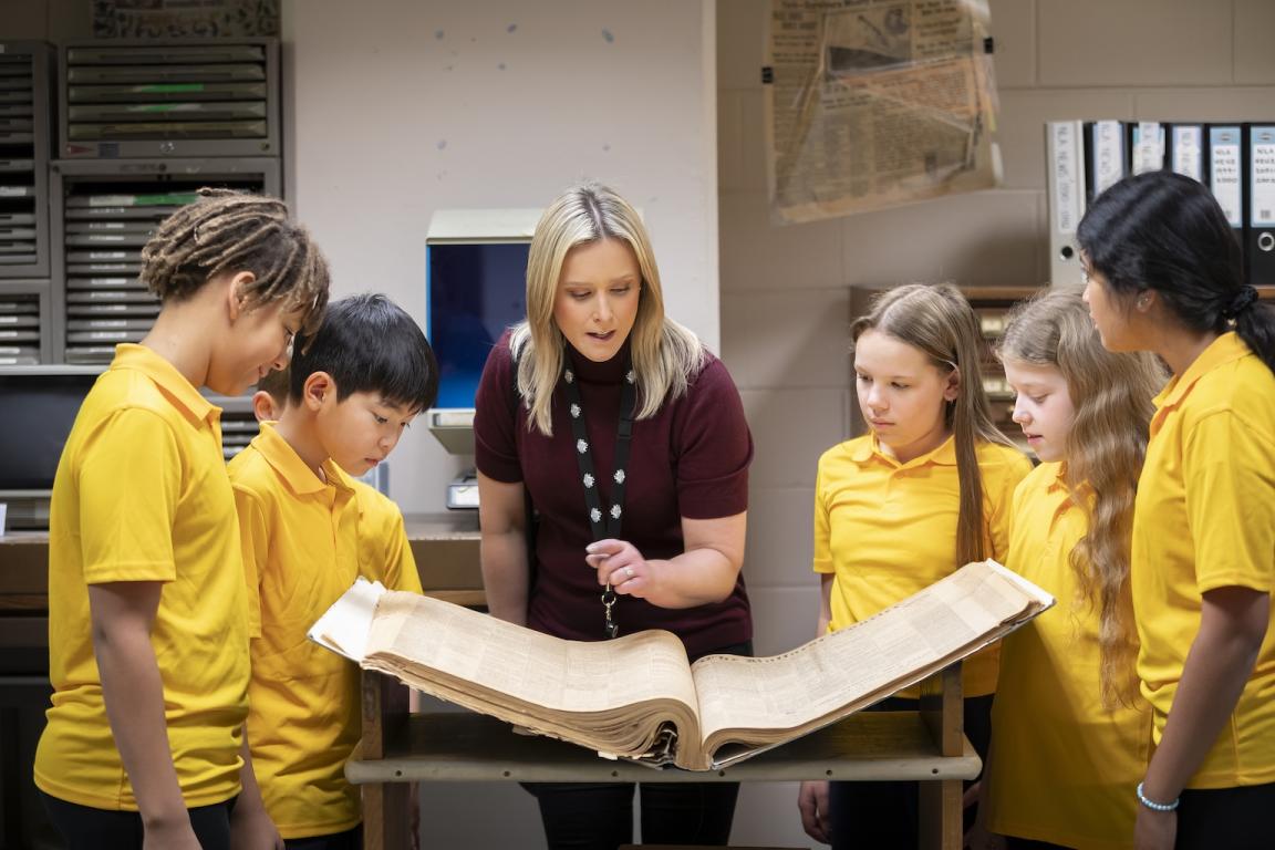 Five young school choldren standing around an old book with a Library staff member explaining the history of the book