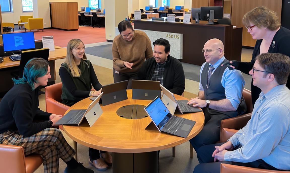 Seven librarians of the National Library sitting or standing around a table talking, smiling and looking at laptops