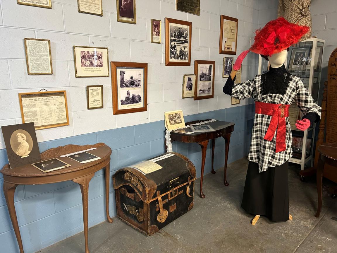 A display of items from the Adelaide Repertory Theatre collection. This includes several small side tables of photographs, a trunk and mannequin wearing a costume displayed in front of a wall of framed material.