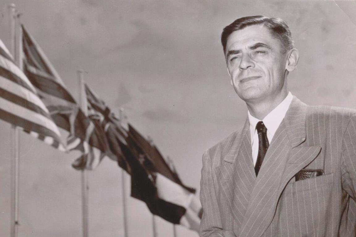 A black and white photograph of a man in a suit standing in front of flagpoles with the US and Australian flags