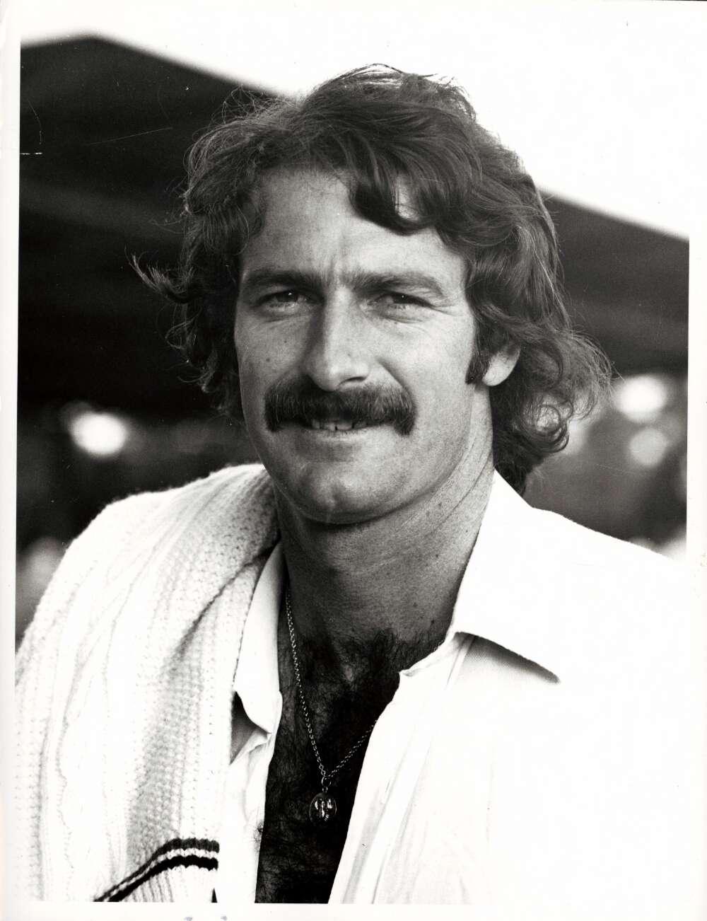 A black and white portrait of Dennis Lillee wearing cricket whites. A jumper is draped over his right shoulder.