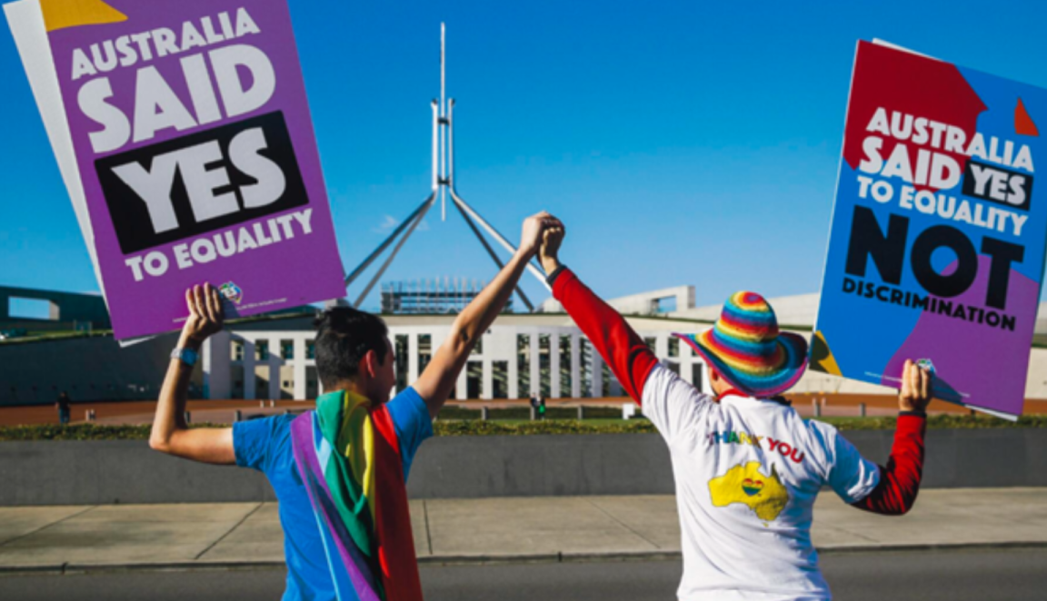 Two men in colourful outfits holding their linked hands up in front of Parliament House. Their other hands hold up signs reading 'Australia said YES to equality' and ''Australia said YES to equality not discrimination' 