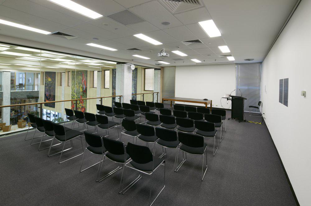 Medium-sized room with rows of chairs facing a large screen. One wall is made of windows which overlook the Library foyer