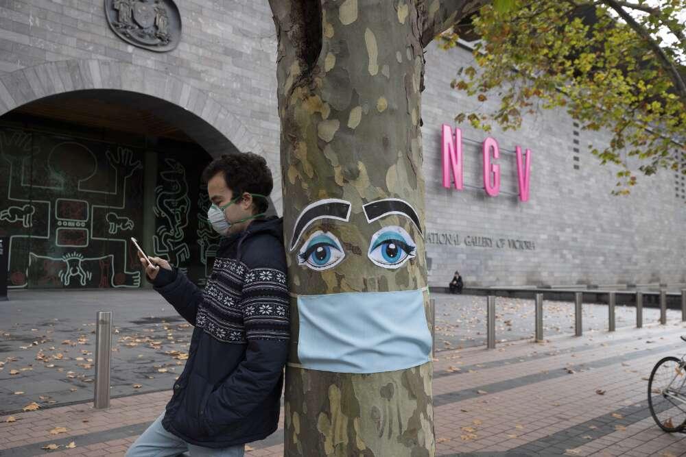 A man wearing a face mask leaning against a tree that is decorated with a face mask and eyes outside the NGV in Melbourne