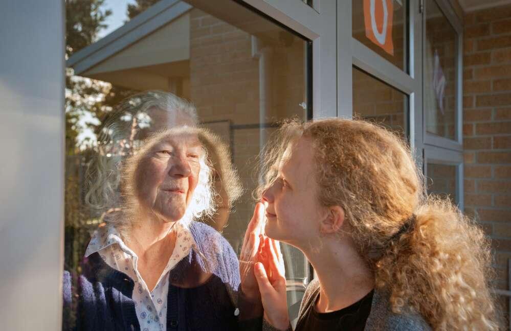 A grandmother and granddaughter separated by a window raise their hands in greeting while looking at each other