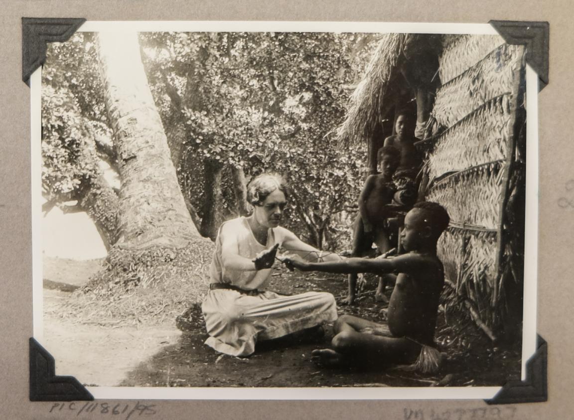 Black and white photo in a photo album showing an older woman, Camilla Wedgwood, playing a game with a child, Oaruaru, both of whom are sitting on the ground