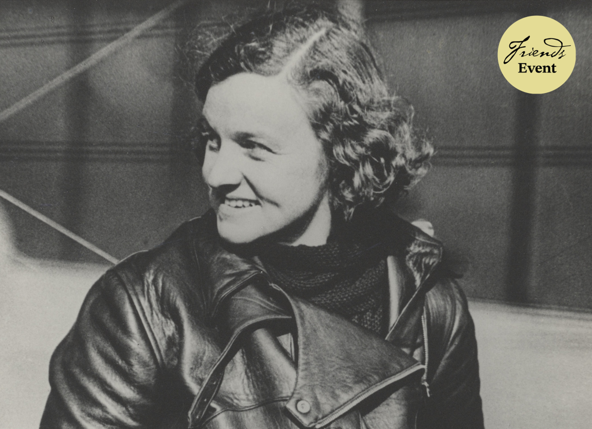 A woman stands in the centre of this image, looking to the left and smiling. She is in front of an aeroplane and wearing a jacket.