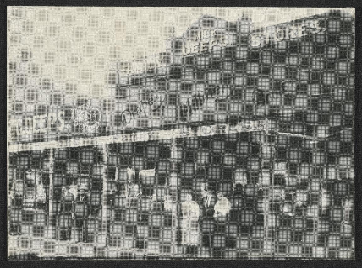 Black and white photograph of the Deeps family shops which sold drapery, millinery, boots and shoes. There are five men and two women standing at the front of the shops under the verandah.