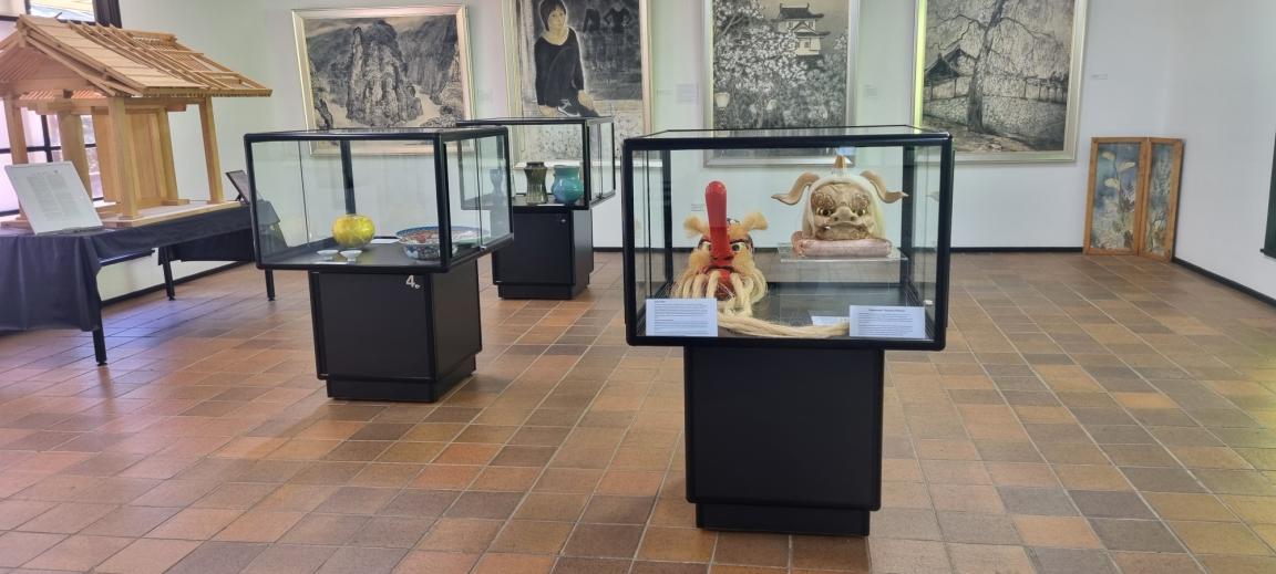 The room of an exhibition gallery. Among other things, four artworks are hanging along one wall, and objects are displayed in glass cases in the foreground.