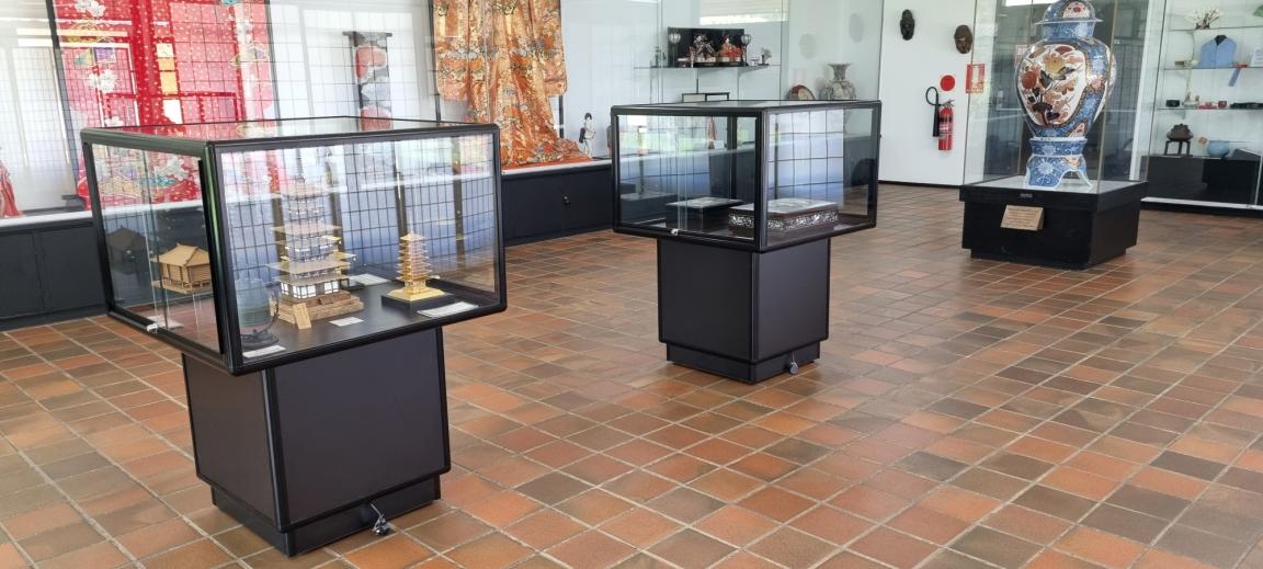 The room of an exhibition gallery. A variety of objects, from garments to dolls, adorn the walls in numerous display cases. A further three display cases are in the foreground of the image.