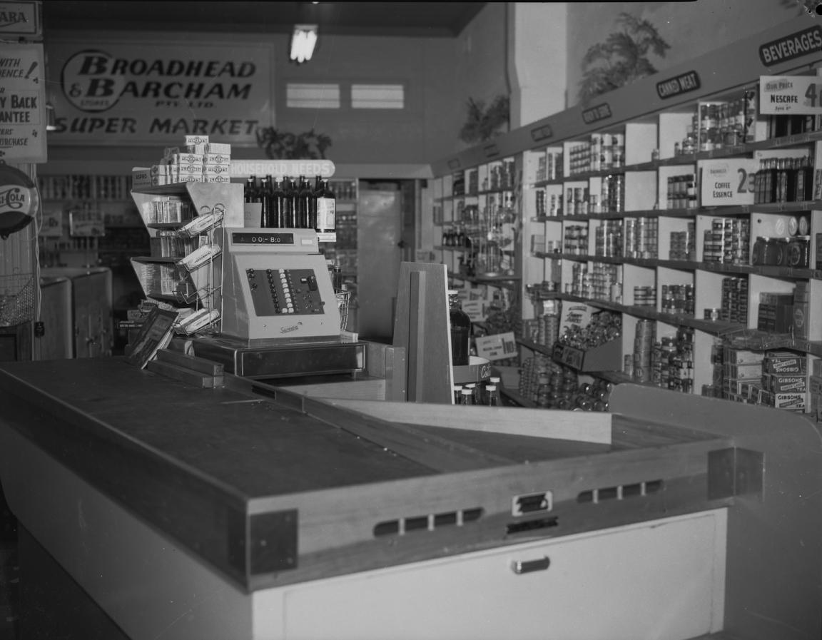 Black and white photo of a supermarket cash register and conveyor belt, with shelves of groceries in the background.