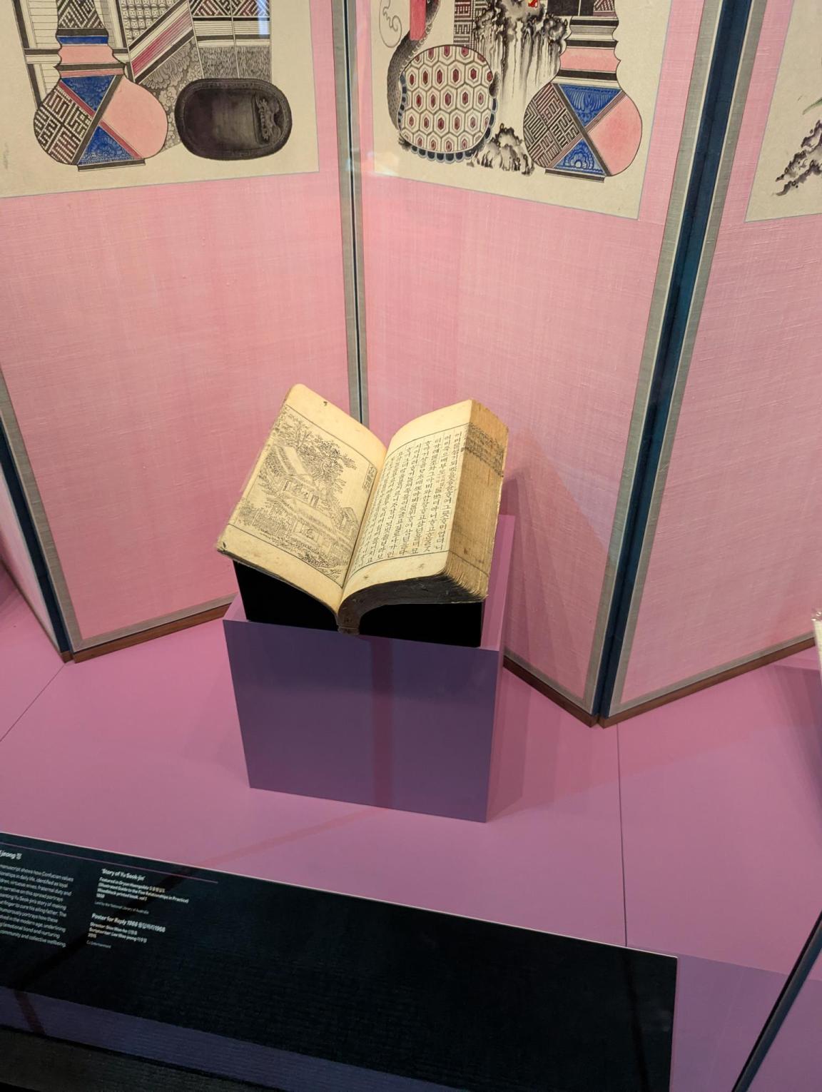 A book lies open on a support structure in an exhibition display case. The surrounding colour and items are pink.