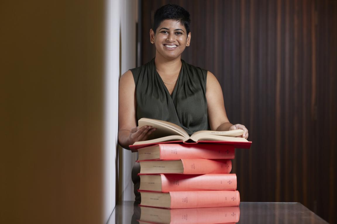 Natasha Naidu standing at a desk behind a stack of books holding one open and smiling at the camera