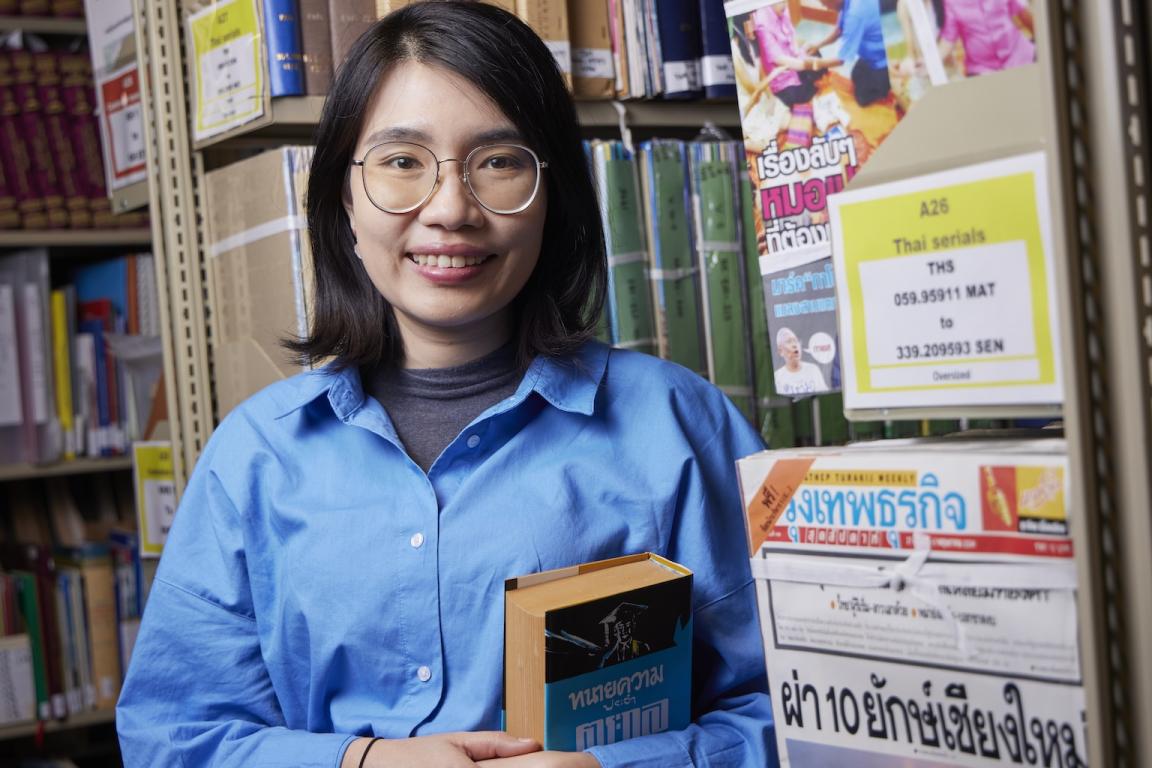 Woman with dark hair, Suprawee (Earn) Asanasak, standing in the stacks of the Library smiling and holding a book