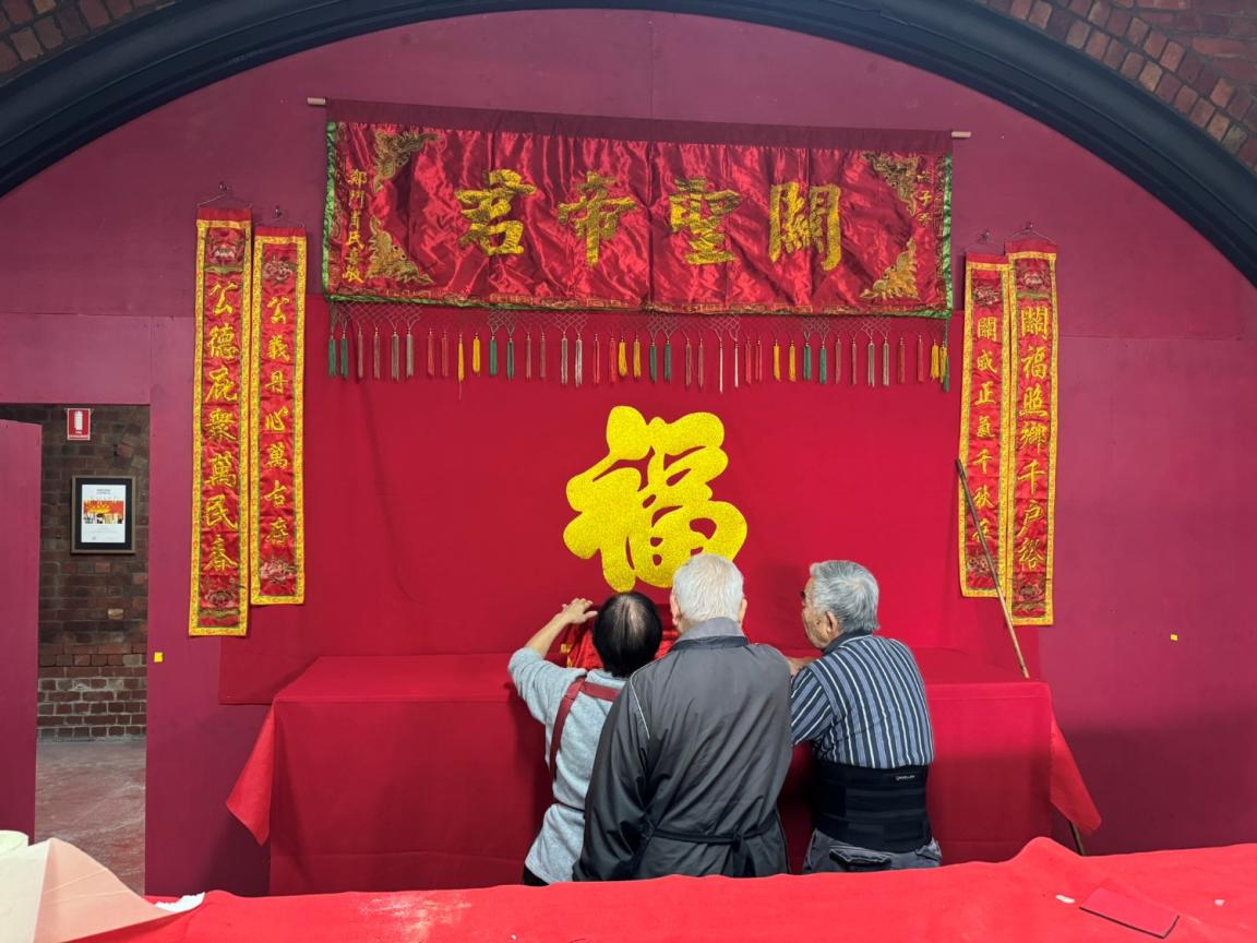 Three people stand in the centre of this image placing items on a temporary structure. The structure is dressed in maroon and red colours with yellow script written on the silk banners. 