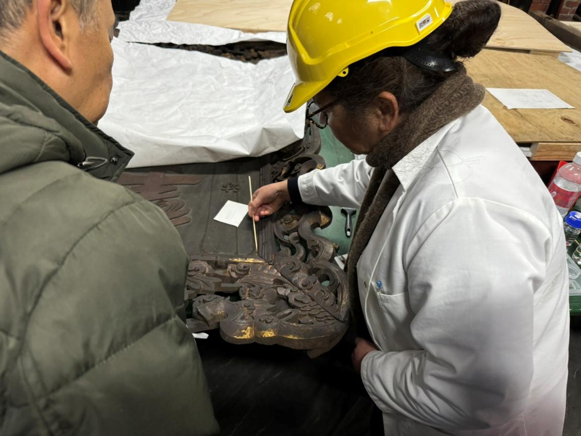 A woman stands on the right-hand side of this image holding a cleaning tool and applying it to a plaque. A man is on the left-hand side watching the cleaning process. 