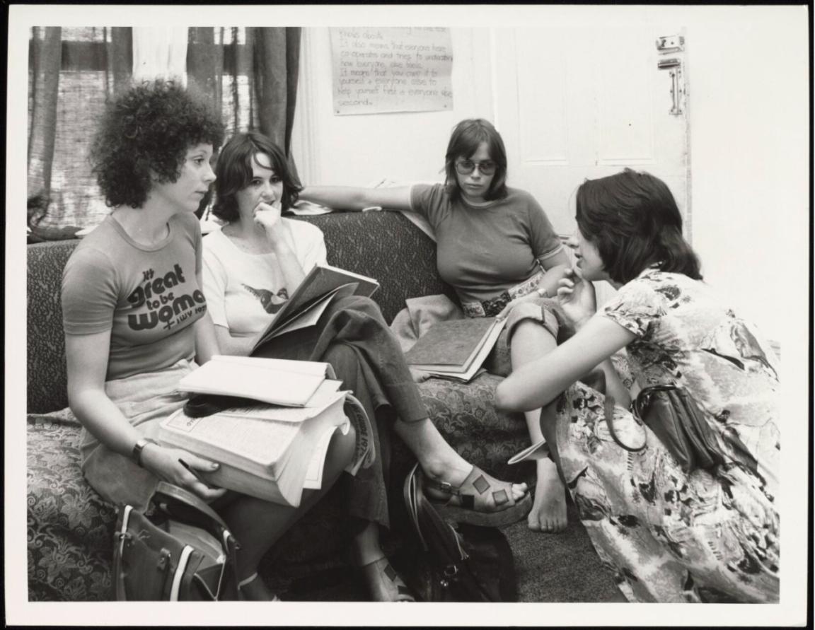 Three women seated on a couch in discussion with one woman crouching on the floor facing the others