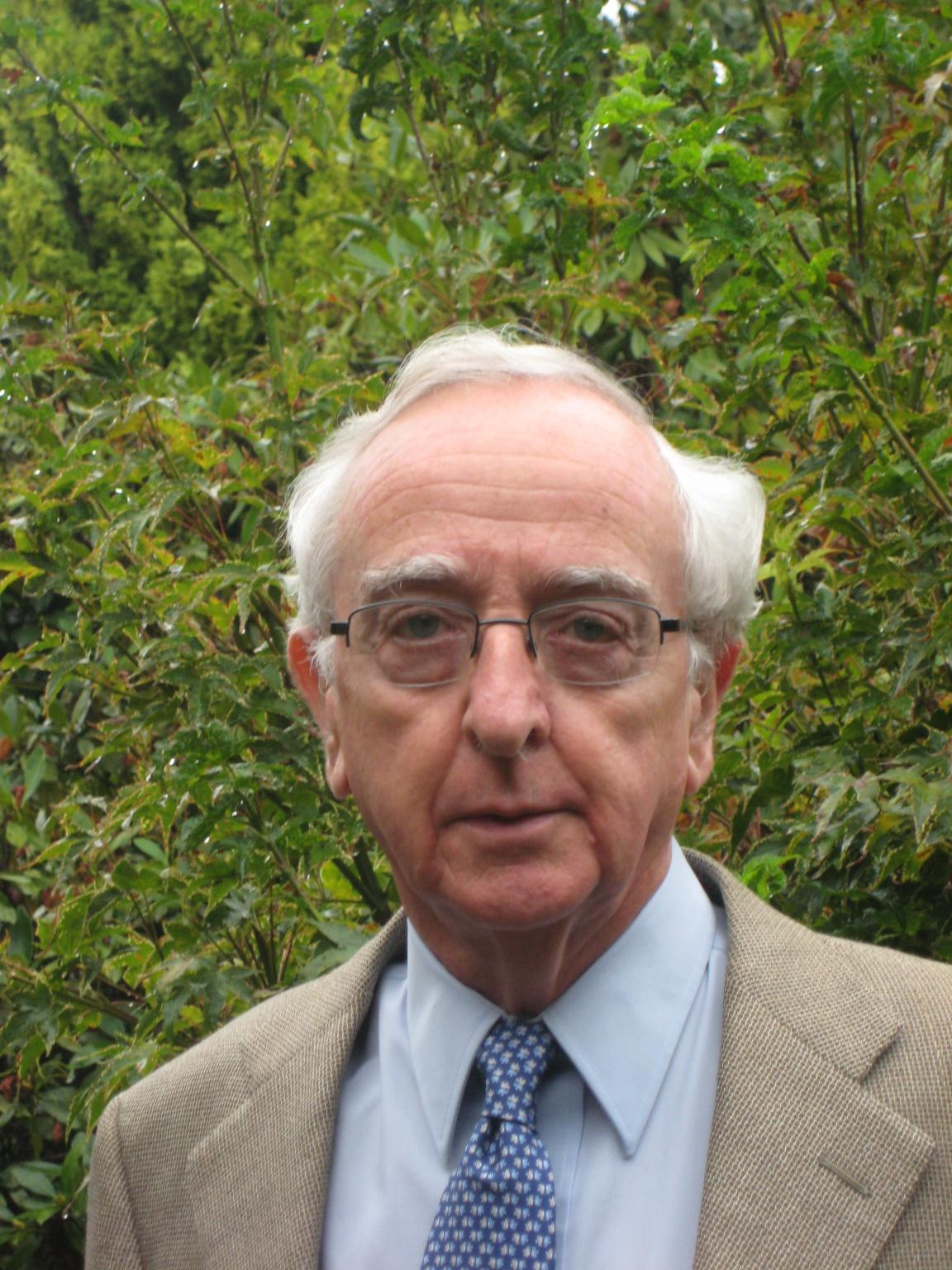 A white haired man wearing glasses, blue shirt and tie and a beige jacket