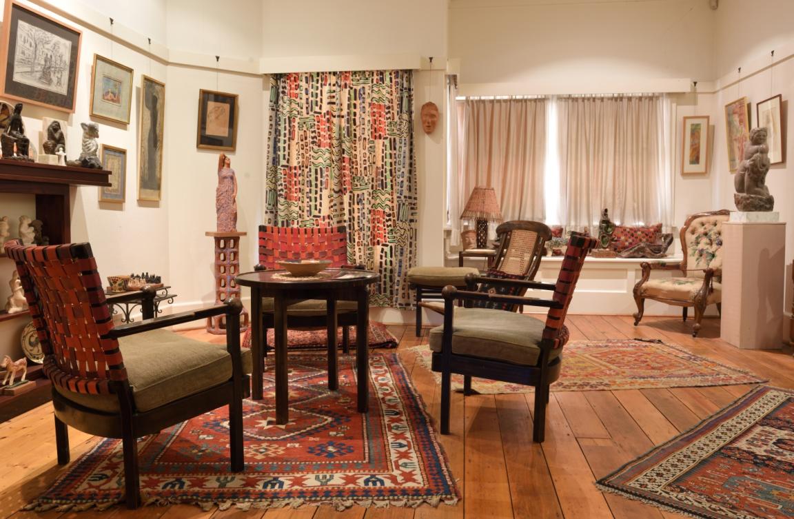 A cosy living room. A small table surrounded by four chairs takes up much of the image. There are a few other seats in the image, and artwork and sculpture adorning the walls and shelves lining the walls.