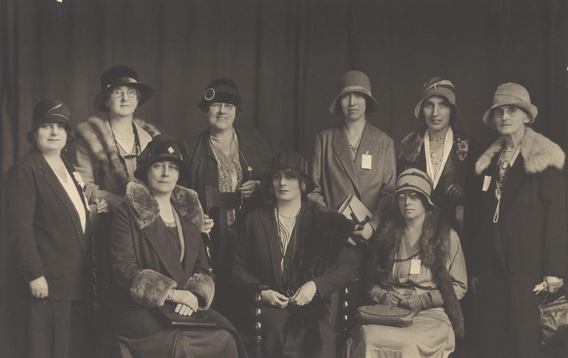 Nine members of the Australian Federation of Women Voters posing for a group photo, some standing and some sitting, in two rows in front of some dark curtains