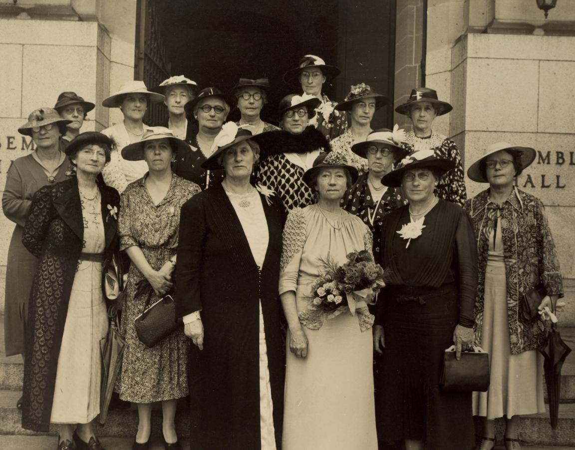 Sepia-toned photo of 16 members of the Australian National Council of Women standing with Dame Maria Ogilvie Gordon on the steps in front of a building