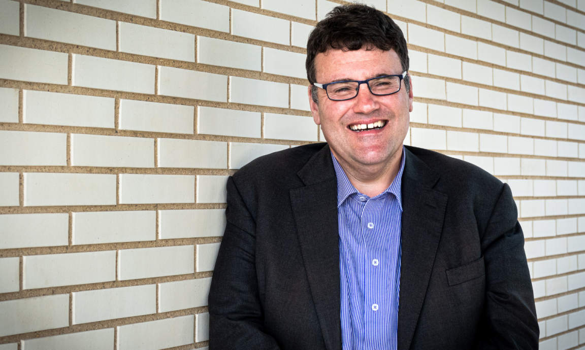 A man wearing a blue button down shirt and blazer smiling at the camera while standing in front of a white brick wall