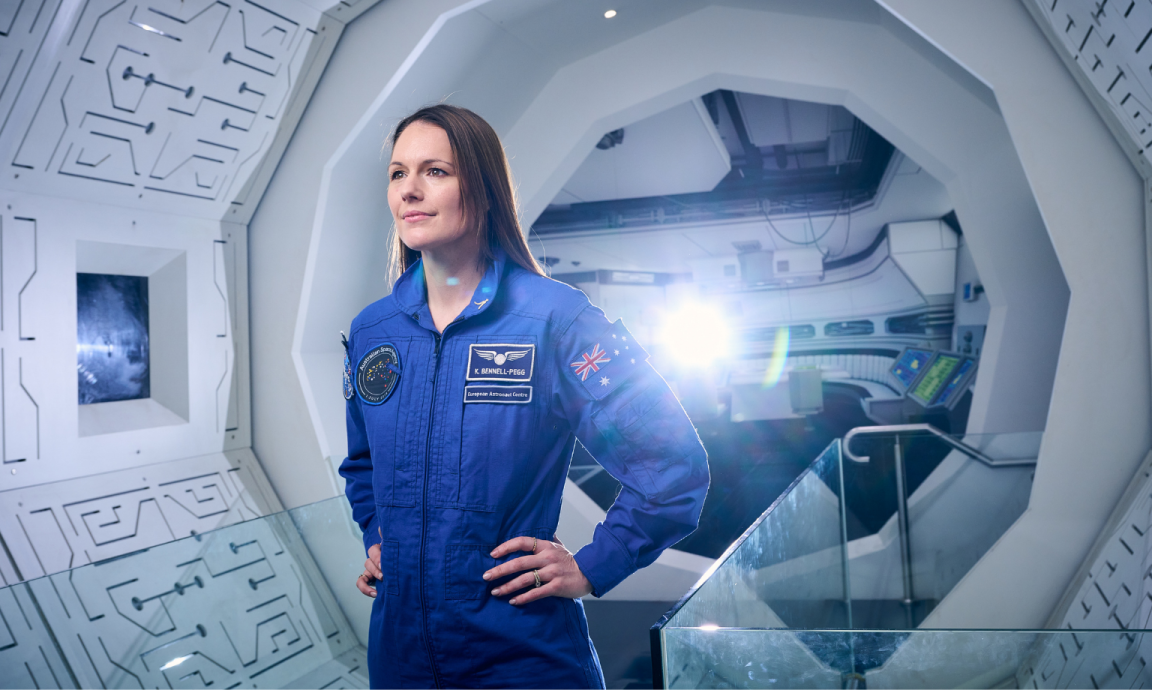 A woman in a space station wearing her astronaut suit with computers behind her