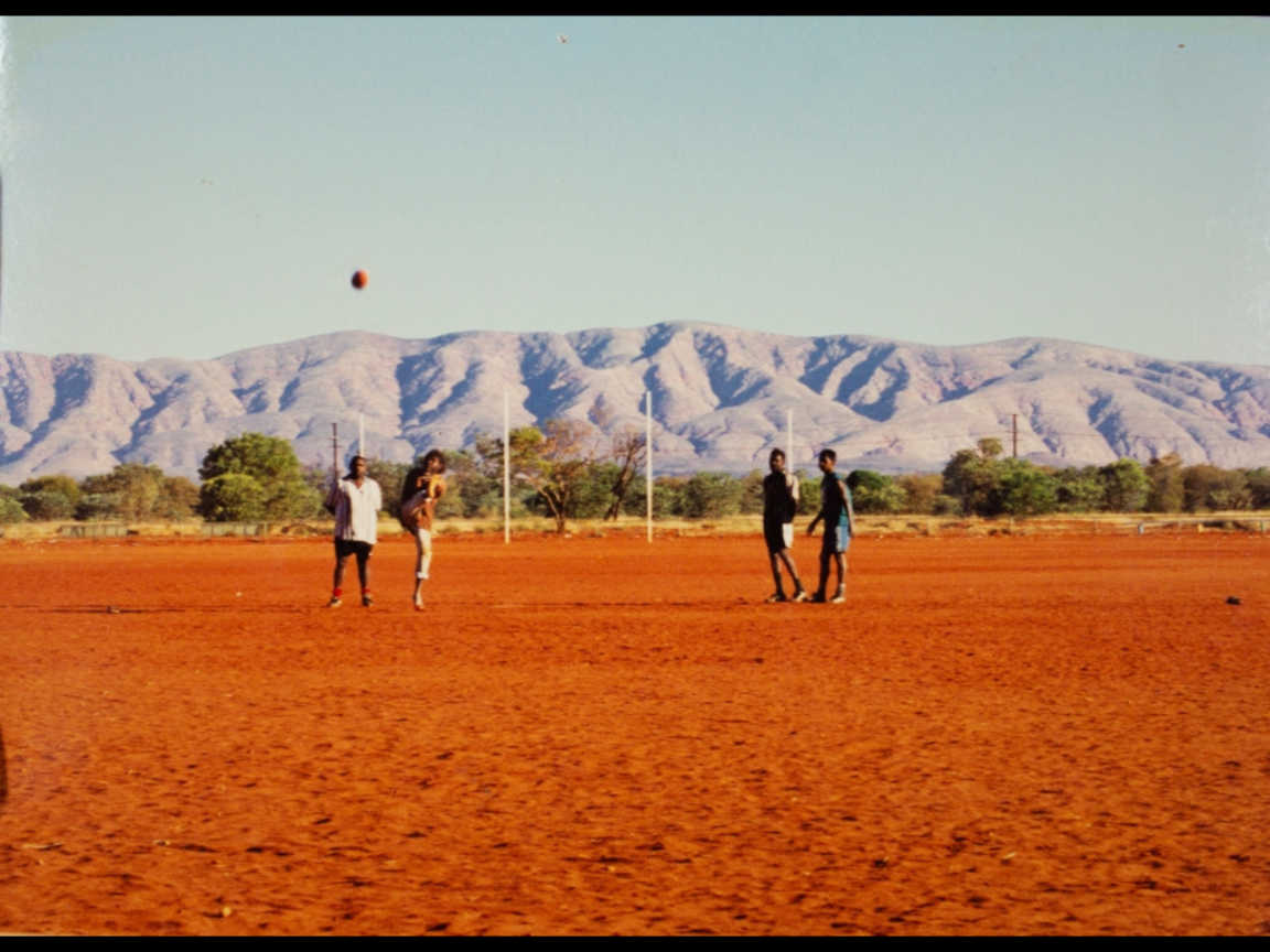Four people play AFL football on a field. There are hills in the background.