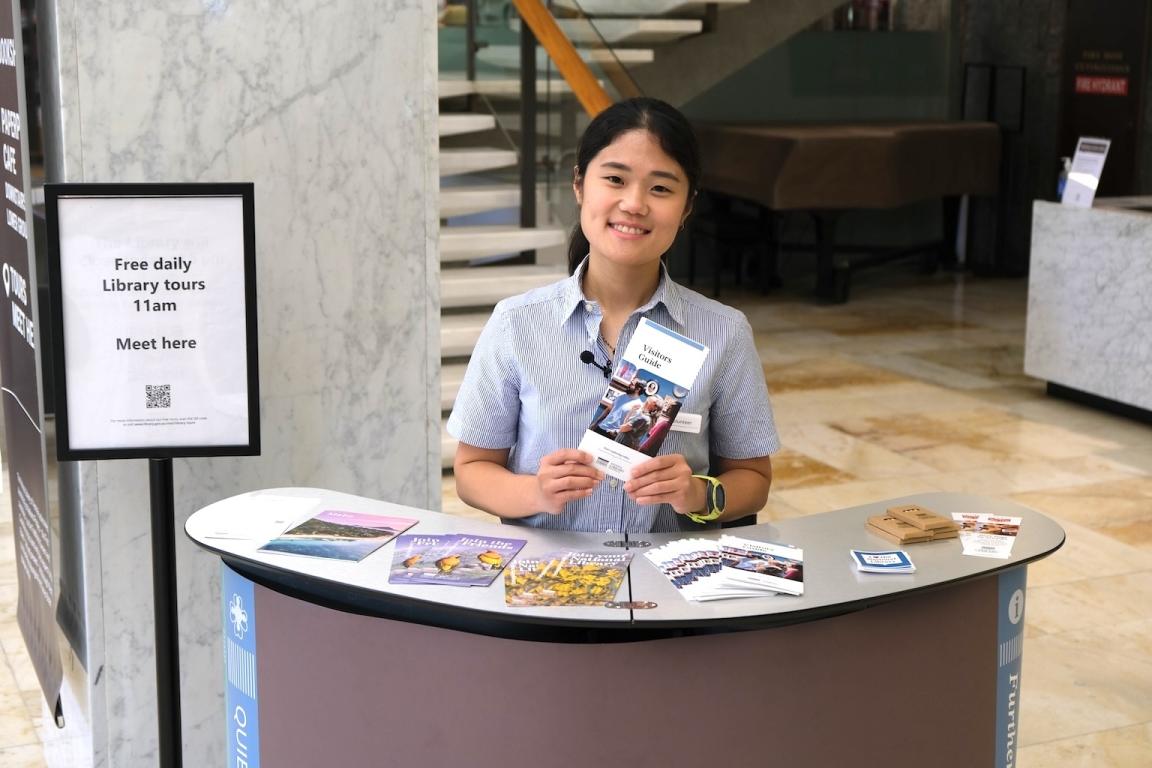 Young Library volunteer standing behind the meet and greet desk smiling and holding up a Visitor Guide brochure