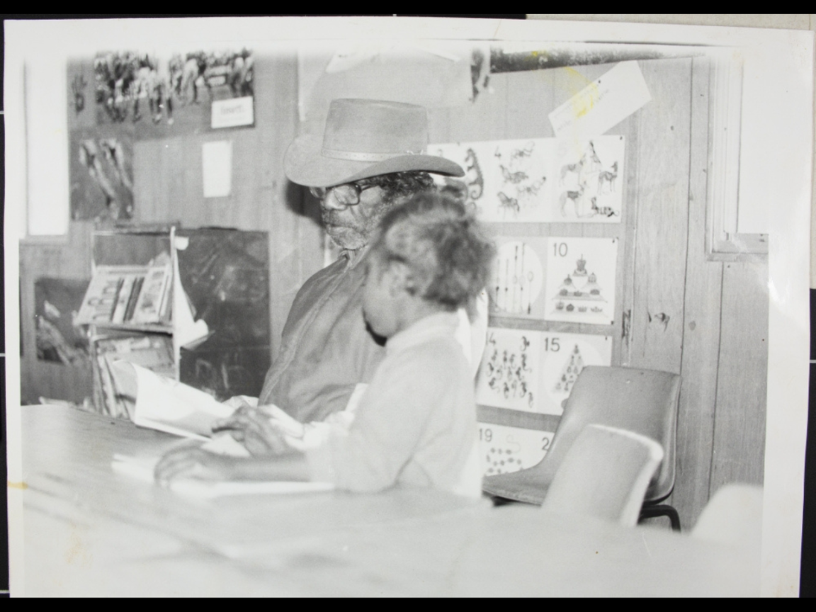 A man and a child sit at a desk in a classroom reading a book.