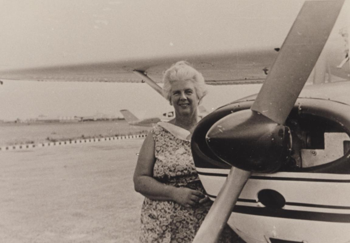 Sepia-toned photo of an older woman in a floral dress standing near the front propeller of a small plaine