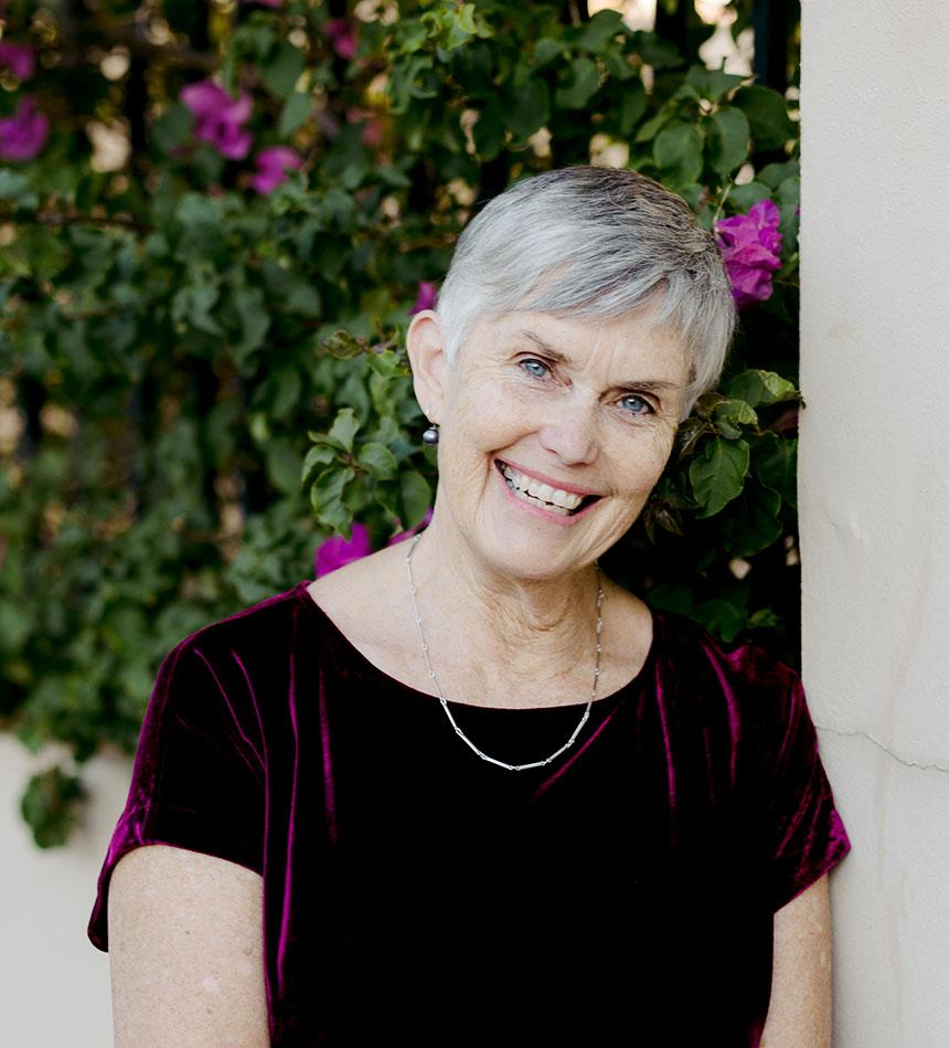 A portrait of Dianne Wolfer. She wears a burgundy dress and a silver chain, and stands infront of a rosebush garden