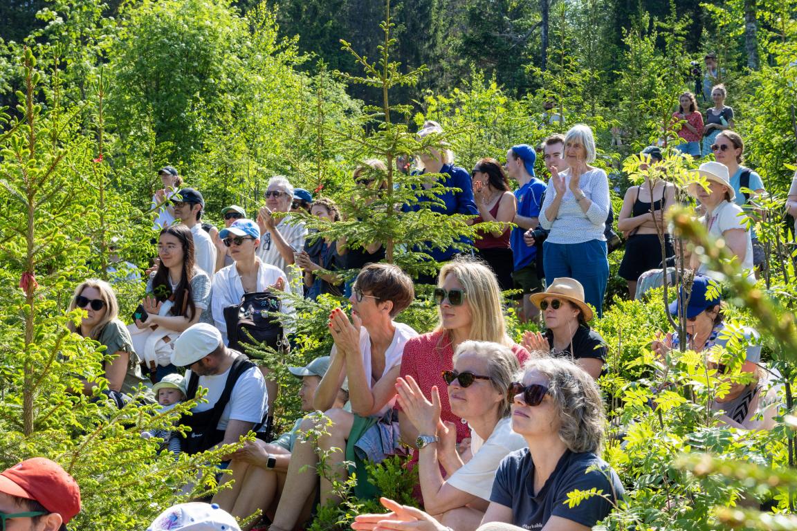A group of people sitting in a forrest, listening to a speech