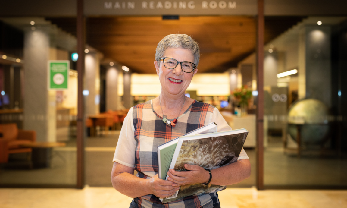 A woman wearing glasses with short hair, holding books in front of the Main Reading Room at the National Library of Australia