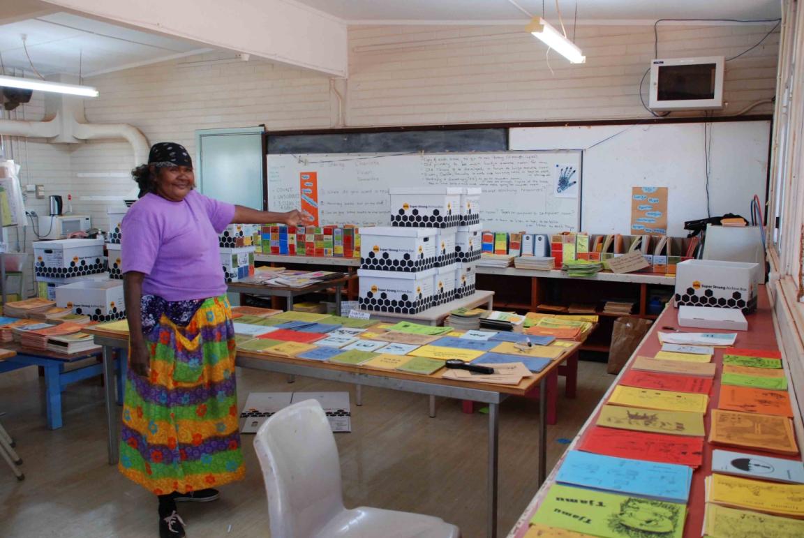 A woman stands in a classroom gesturing at piles of readers laid out on the tables in the classroom.