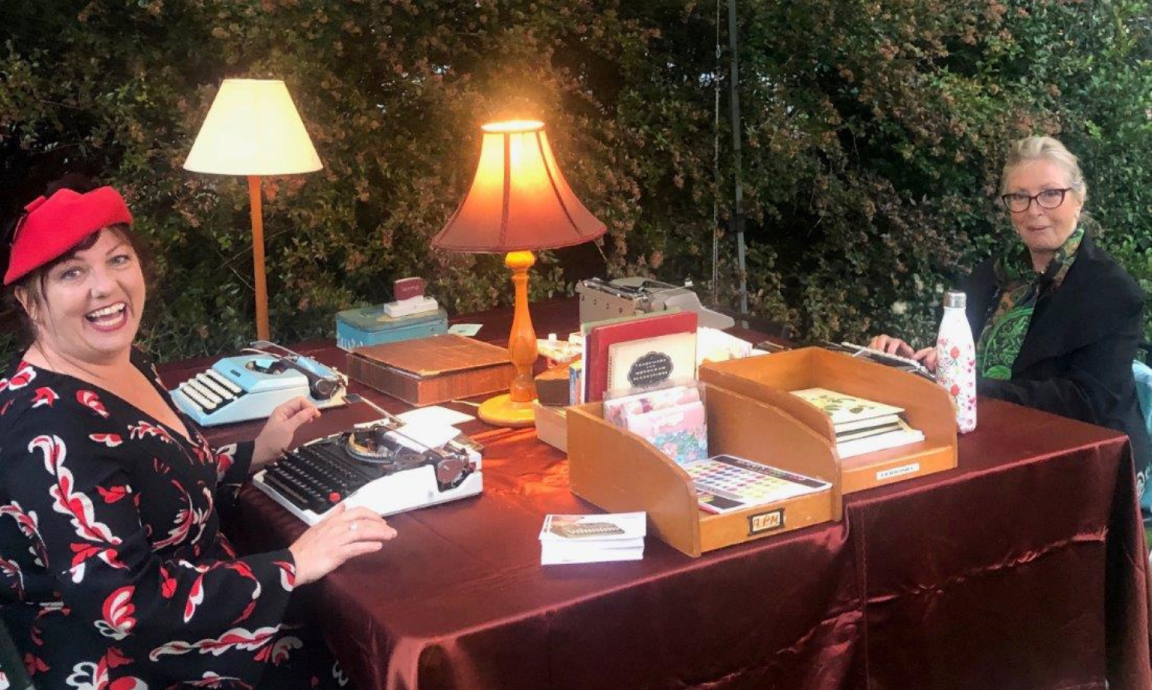 Two women are seated at a table covered in red cloth and laid with vintage lamps, stationery and typewriters