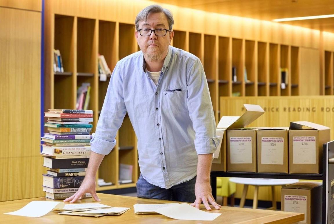 A man in a blue shirt leaning over a desk with papers and books