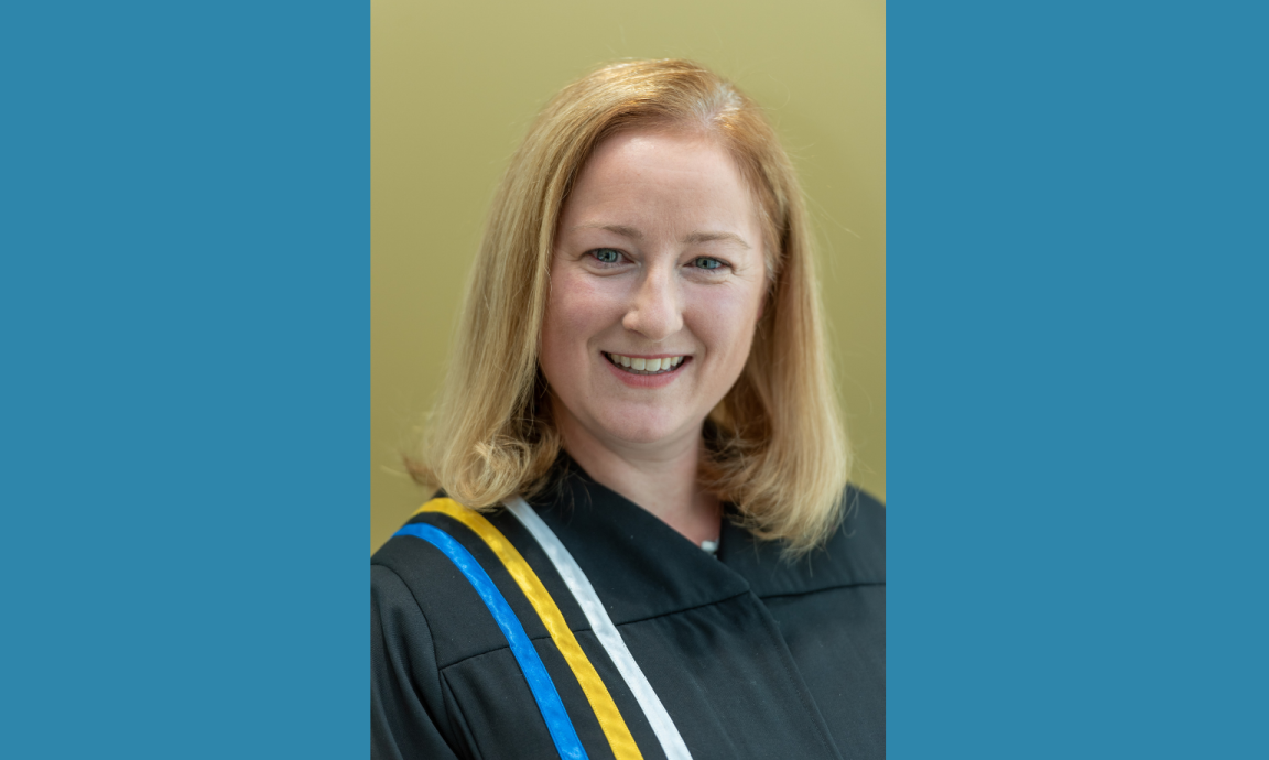 A blond-haired woman wearing courtroom robes with blue, yellow and white ribbons on the right shoulder