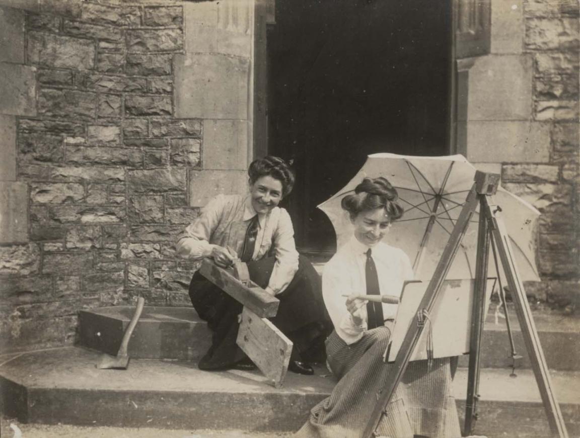 Two women sitting on a porch, on holding an umbrella and painting at an easel, the other holding woodworking tools