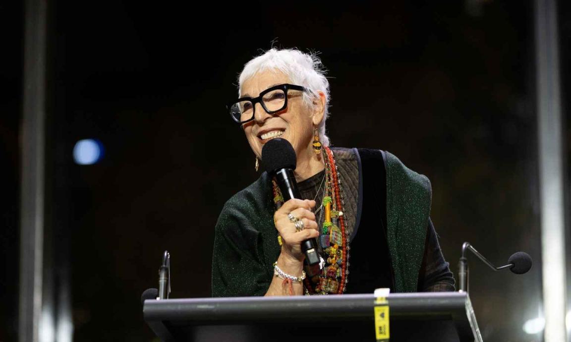 A woman with white hair and dark-rimmed glasses is speaking into a microphone