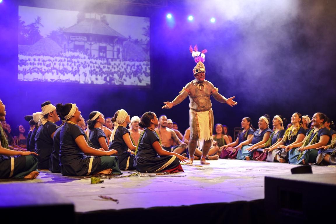 Several dancers on their floor with their legs crossed looking up at another dancer walking between them with his arms out