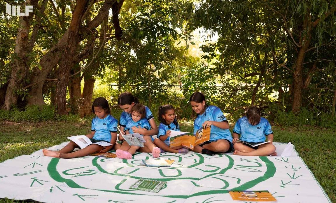 A group of young girls are sitting on a large white and green mat outdoors, reading books together in a sunny, lush green forest setting.