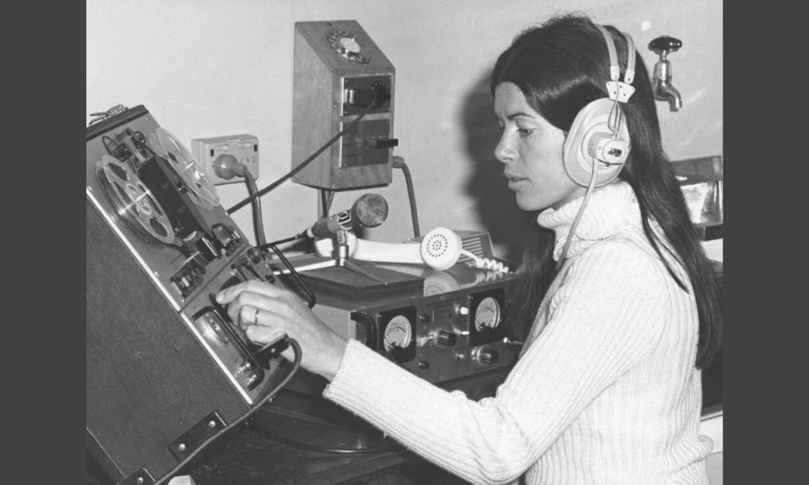 A black and white photo of a woman wearing headphones, operating a vintage audio recording or mixing console in a studio setting.