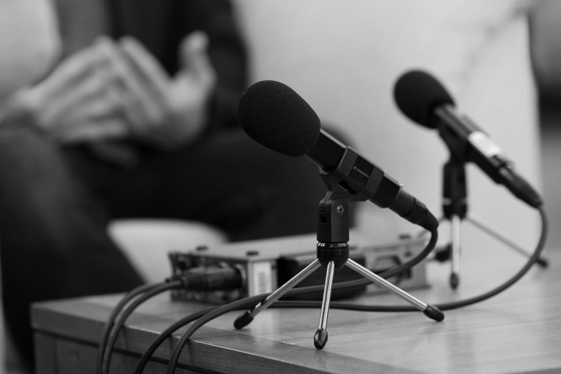Black and white image of recording equipment and microphone on desk, with a pair of hands in the background