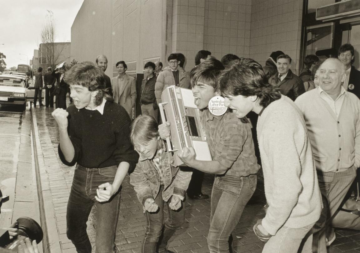 Black and white photo of four teenage boys, one holding a boombox, singing or yelling into a large camera