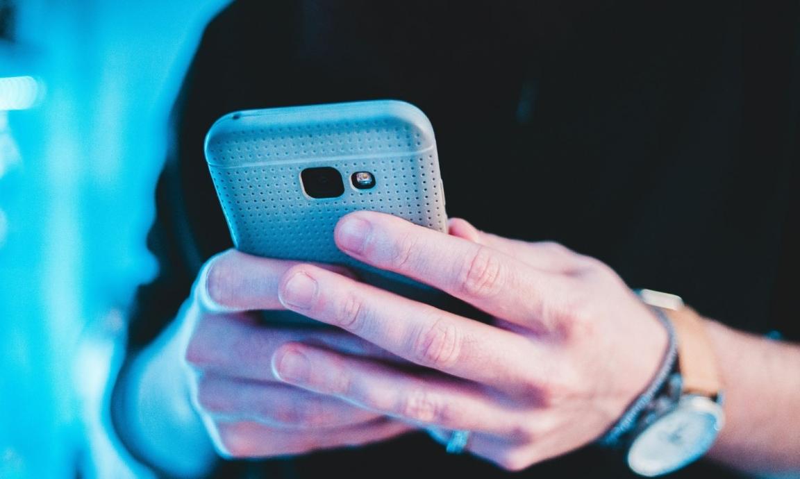 Hands of a person holding a light blue smartphone in their hands against a dark background.