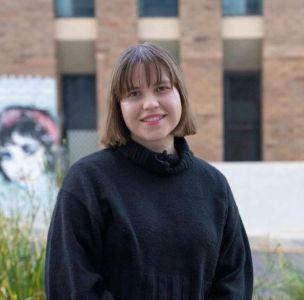 A young woman with straight brown hair and bangs wearing a black turtleneck sweater, standing outdoors in front of a modern brick building.