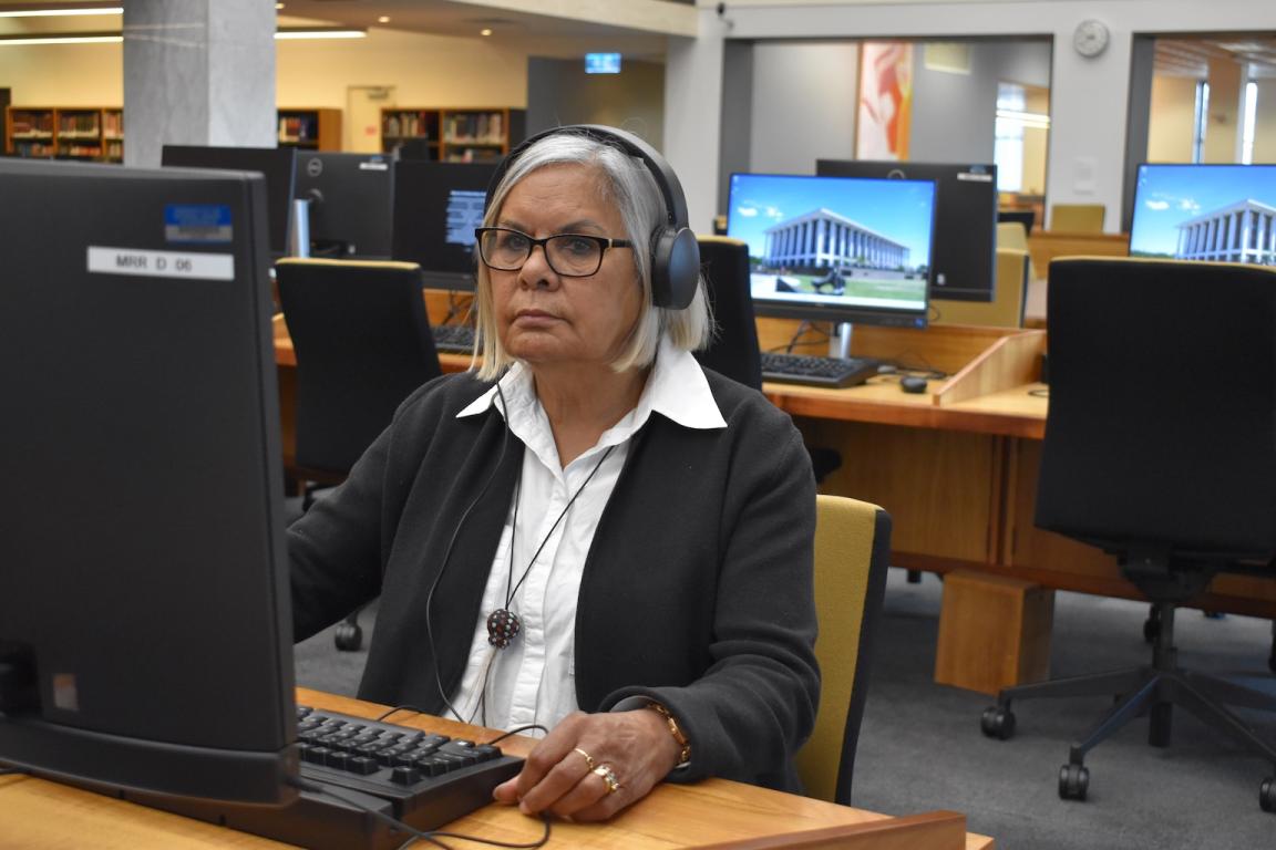 Wirangu Elder Sandra Miller sitting at a computer in the Main Reading Room wearing headphones and listening to an oral history recording